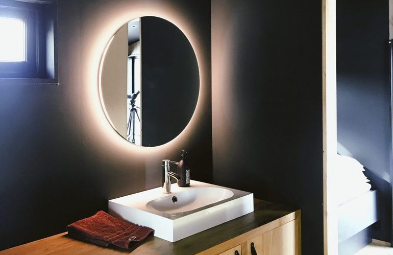 Okrúhle zrkadlo s podsvietením v kúpeľni s tmavými stenami.